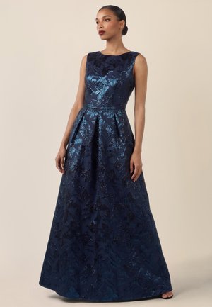 Frau trägt ein ärmelloses, marineblaues, floral gemustertes Abendkleid und steht vor einem schlichten beigen Hintergrund.