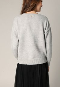 Pull en maille gris clair avec une coupe décontractée, ourlet côtelé et un petit bouton décoratif au dos. Associé à une jupe plissée noire.