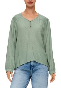 Lichtgroene blouse met lange mouwen, een V-hals, knoopdetails, textuurstof en gerimpelde schouderaccenten, gedragen over blauwe spijkerbroeken.