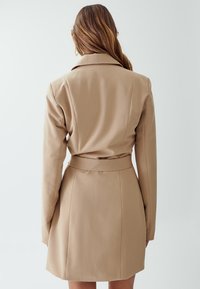 Beige trench coat dengan sabuk, lengan panjang, potongan yang pas, dan tekstur yang halus. Panjang hingga lutut dengan kerah berpatah dan detail ventilasi di belakang.