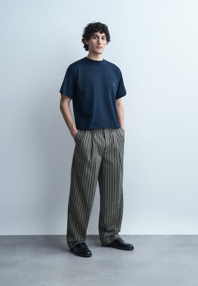 Νεαρός άνδρας με σγουρά μαλλιά, που φοράει μπλε σκούρο μπλουζάκι, φαρδιά παντελόνια με ρίγες και μαύρα παπούτσια, στέκεται με τα χέρια στις τσέπες.