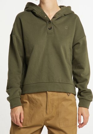 Personne portant un pull à capuche vert olive avec patte de boutonnage et pantalon beige, debout les bras détendus le long du corps.