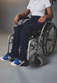 Ratiņkrēsls ar metāla rāmi. Lietotājs valkā tumši zilos bikses un zilos sporta apavus ar baltām akcentiem. Gluda grīda, neitrāla pelēka fona.