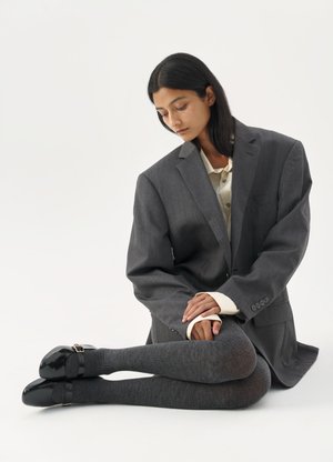 Moteris sėdi ant grindų, dėvi per didelį pilką švarką, smėlio spalvos marškinius, pilkas pėdkelnes ir juodus Mary Jane batus, žiūri žemyn susikibus rankomis.