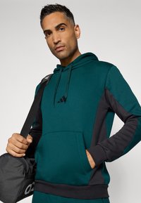 Zeleno-modra kapucha s kapuco iz mehkih materialov, ki ima sprednji žep, vrvici in logotip adidas na prsih.