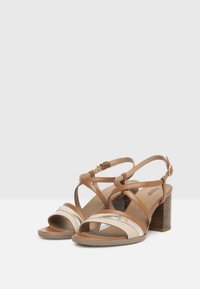 Braune und cremefarbene Ledersandalen mit Blockabsatz, ausgestattet mit Kreuzriemen und einem metallischen Akzent, offenes Zehendesign und Schnalle am Knöchel.