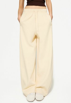 Persoon draagt brede, crèmekleurige sweatpants met zijzakken, gecombineerd met witte sneakers en een donkeren cropped top.