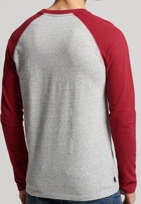 Man i en långärmad raglantröja med grå kropp och mörkröda ärmar, stående vänd bort.