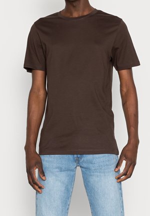 Homme portant un t-shirt marron uni à manches courtes et un jean bleu clair sur fond blanc.