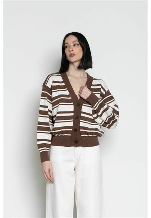 Donna con capelli neri taglio bob, che indossa un cardigan a righe marrone e bianco e pantaloni bianchi larghi su uno sfondo chiaro uniforme.