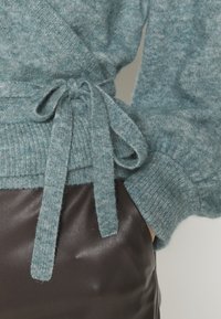 Turkos stickad tröja med en mjuk textur, med en knytning vid midjan och ribbade manschetter mot en mörk läderbakgrund.