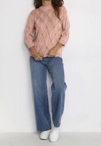 Hellrosa gestrickter Pullover mit Rautenmuster, gerippten Bündchen und hohem Kragen, kombiniert mit lässigen blauen Jeans und weißen Turnschuhen.