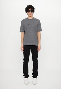 T-shirt cinza riscado com mangas curtas e logo preto, combinado com jeans pretos. Ténis brancos e óculos de sol escuros completam o visual.
