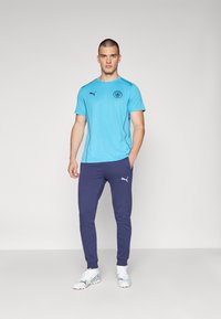 Gaiši zila sporta t-kreksa ar melnu logo un plecu akcentiem, apvienota ar tumši ziliem sporta biksēm, kurām ir balta zīmola apzīmējums un pieguļoši piedurkņu gali.