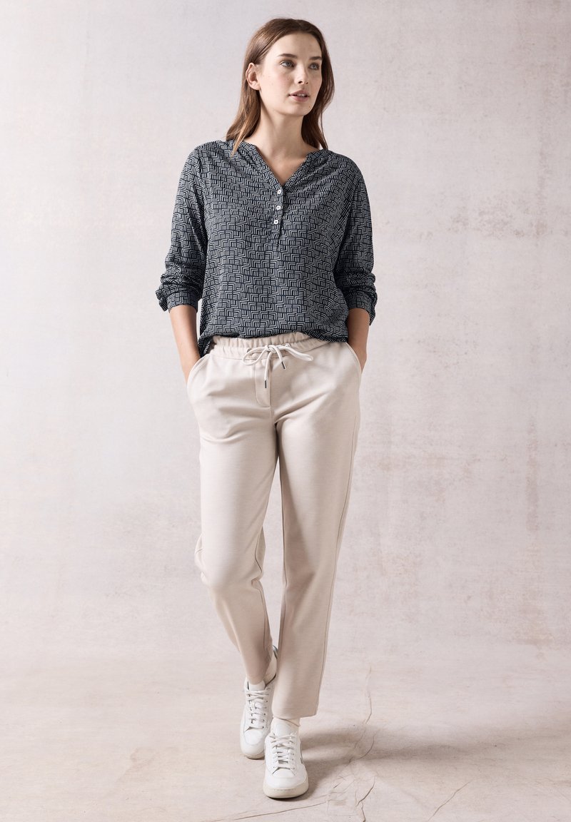 Camicia a maniche lunghe con motivo geometrico bianco e nero, abbinata a pantaloni beigi con cordoncino e sneakers bianche. Tessuto morbido, vestibilità comoda.