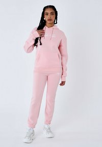 Sudadera rosa con bolsillo tipo canguro y logo, combinada con pantalones joggers rosas a juego. Tejido suave, corte relajado, puños elásticos y cinturilla elástica.