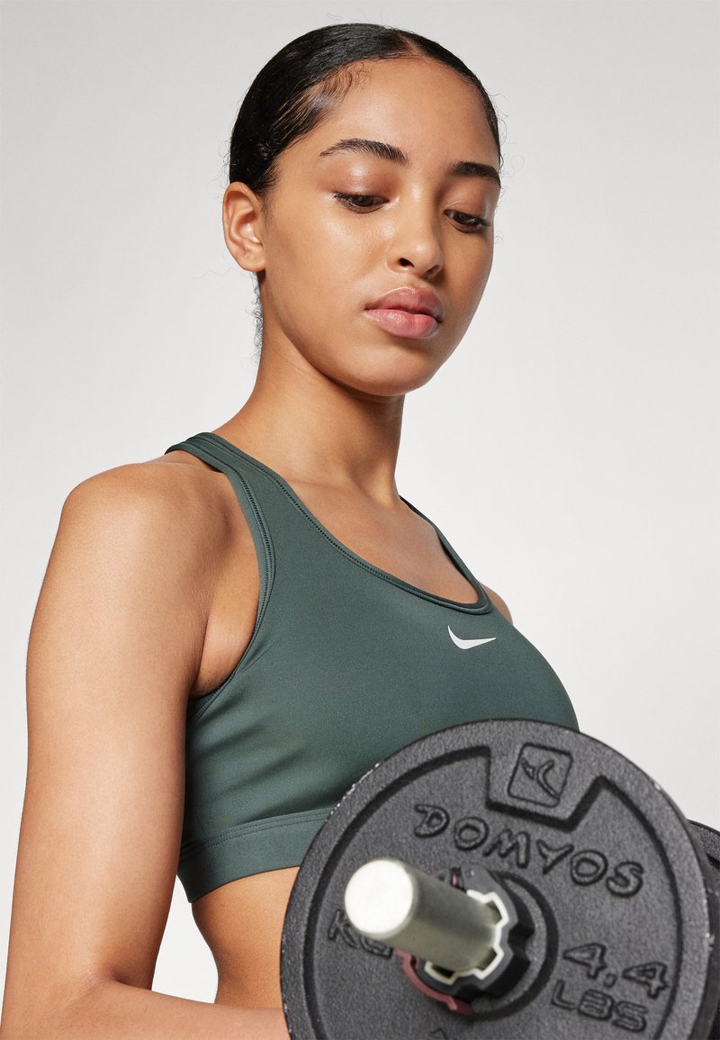 Cropped top sportivo verde con logo bianco, mentre tiene un disco di peso nero con lettere in rilievo. Texture liscia e design aderente.