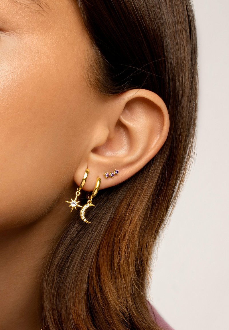Pendientes de oro en forma de aro con charms de estrella y luna creciente, que presentan gemas claras y moradas. Acabado suave y diseño colgante.