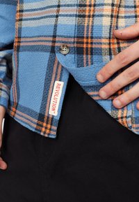 Geruite blouse in blauw, oranje en zwart; geweven stof met textuur; beschikt over een knoopsluiting en een "REVOLUTION" label aan de zoom.