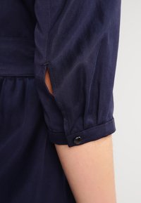 Námořnická modrá košile s rukávem, který má na lokti nařasenou látku, se zapínáním na knoflík na manžetě a bočním otvorem blízko ramene.