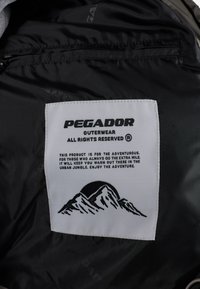 Svart yttertøy med en hvit etikett som leser "PEGADOR," med fjellgrafikk og tekst om eventyr. Glidelåsdetalj synlig.
