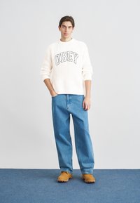 Pulover alb tricotat cu textul „OBEY”, blugi largi albaștri și pantofi maro slip-on. Modelul stă în fața unui fundal neutru.