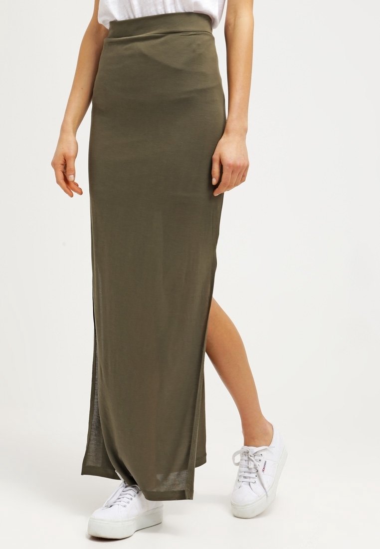 Femme portant une jupe longue vert olive avec une fente latérale et des baskets blanches, debout devant un fond clair et uni.