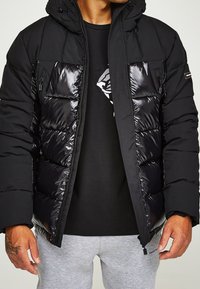 Svart pufferjacka med glansig yta, med dragkedjefickor och huva. Bärs över en svart skjorta och grå sweatpants.