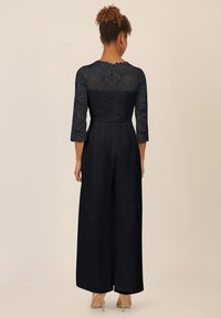 Zwart jumpsuit met een kanten lijfje met bloemenpatronen, driekwart mouwen en een wijde pijp; afgewerkt met een achterrits.