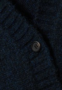 Marineblauer, strukturierter Strickcardigan mit einem auffälligen schwarzen Knopf und sichtbaren Fasern, der ein dickes, gemütliches Material präsentiert.