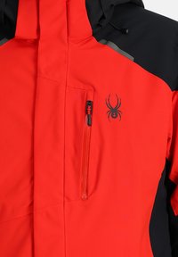 Rote und schwarze Skijacke mit einer wasserdichten Außenschicht, einer vorderen Reißverschlusstasche und einem Spinnenlogo auf der Brust. Mit Kapuze und strukturiertem Finish.