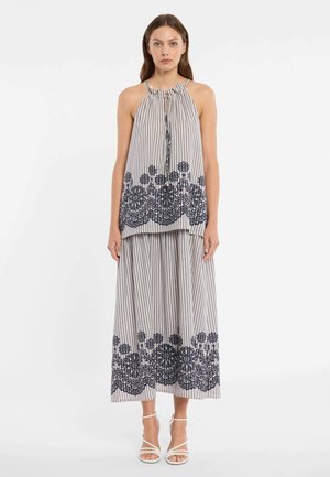 Femme portant un haut dos nu rayé à col halter et une jupe large assortie avec une broderie florale noire le long de l'ourlet, associée à des sandales blanches.