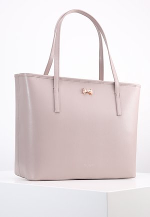 Beige tekstiiliton nahka käsilaukku, jossa on kaksi kahvaa. Edessä pieni metallinen nauha koristeeksi ja teksturoitu pinta. Neliömäinen muoto.