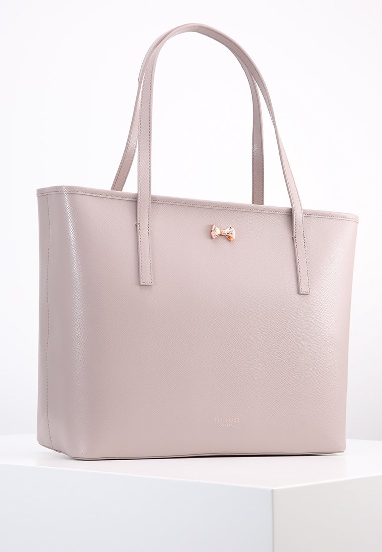 Beżowa torebka z sztucznej skóry z dwoma uchwytami. Posiada mały metaliczny akcent w kształcie kokardy z przodu oraz teksturowaną powierzchnię. Kwadratowy kształt.