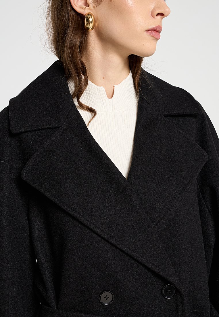 Czarny wełniany płaszcz o podwójnym zapięciu, z szerokimi klapami i dwoma guzikami. Pod płaszczem znajduje się teksturowany biały sweter.