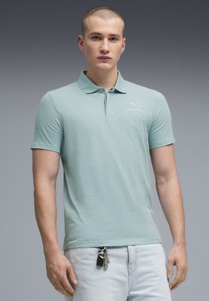 Giovane uomo con capelli corti che indossa una polo McLaren azzurro chiaro e jeans chiari con un portachiavi appeso all'asola della cintura.