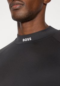 Czarny top sportowy z wysokim kołnierzem i białym logo "BOSS" z przodu. Gładka faktura i dopasowany krój.