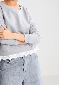 Ljusgrå sweatshirt med vit spetskant i nederkant, som har en avslappnad passform och långa ärmar. I kombination med ljusblå denimjeans.