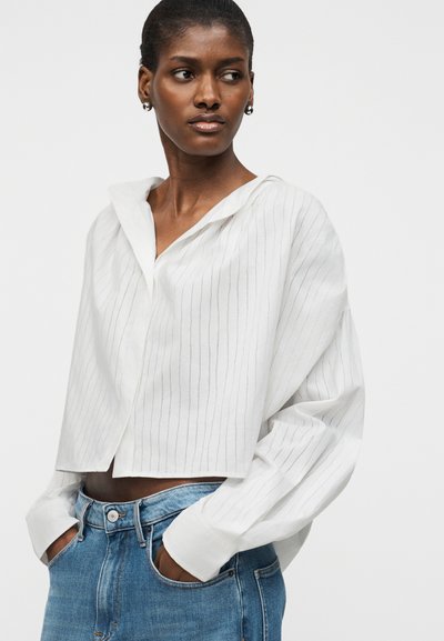 Γυναίκα που φοράει λευκό, μακρυμάνικο, κοντό μπλουζάκι με λεπτές κάθετες ρίγες και μπλε ψηλόμεσα τζιν, με τα χέρια στις τσέπες, κοιτάζοντας προς τα πλάγια.