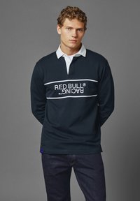 Námornícky dlouhý rukáv polo s bílým límečkem, na přední straně je vodorovný bílý text "RED BULL RACING" přes hrudník. Měkká látka, volný střih.