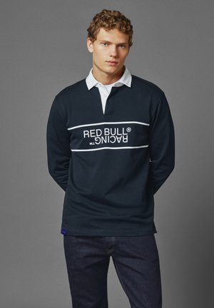 Granatowa koszula polo z długim rękawem i białym kołnierzykiem, z poziomym białym napisem "RED BULL RACING" na piersi. Miękka tkanina, swobodny krój.