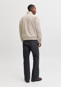 Veste beige à col montant, associée à un jean en denim foncé. La veste présente une texture lisse et des poignets côtelés, avec des détails de coutures.