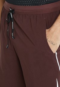 Pantalones cortos deportivos de color marrón oscuro con cintura ajustable y una mano parcialmente en el bolsillo derecho, además de un tatuaje tenue en la muñeca.