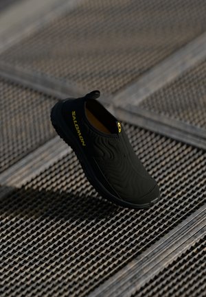 Scarpa nera slip-on con superficie testurizzata, caratterizzata da un logo giallo e un robusto tirante. La suola è progettata per garantire trazione.