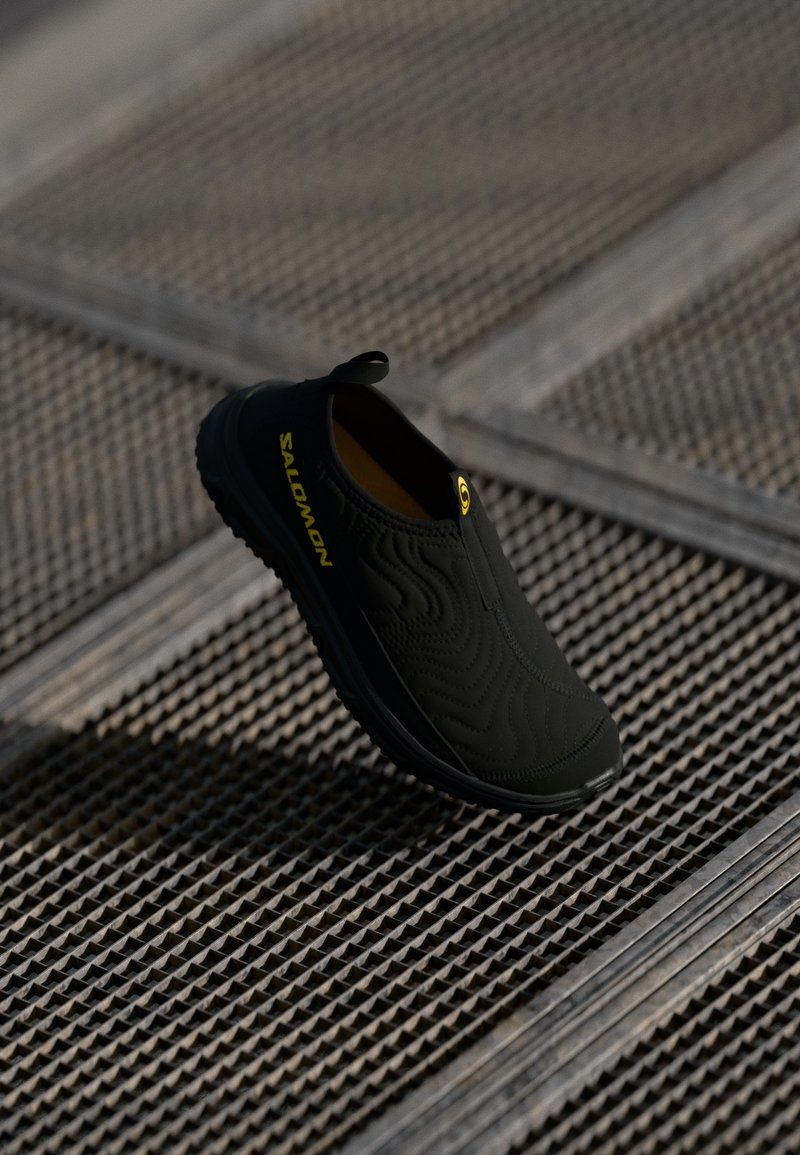 Black slip-on shoe with textured surface, featuring a yellow logo and a pull tab. The sole is designed for traction.