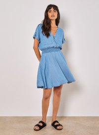 Vestido de mezclilla azul claro con mangas cortas, escote en V y cintura fruncida. La falda fluye hasta mitad del muslo, complementada con sandalias negras tipo slide.