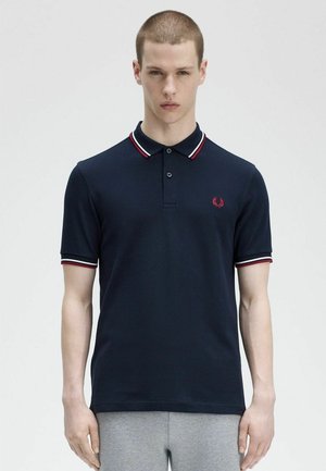 Joven con cabello corto que lleva una camiseta polo azul marino con detalles en rojo y blanco, posando frente a un fondo claro y uniforme.