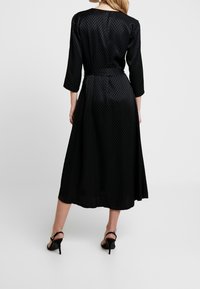 Abito midi nero con un motivo a diamante testurizzato, maniche a tre quarti, vita con cintura e gonna svasata. Indossato con tacchi neri.