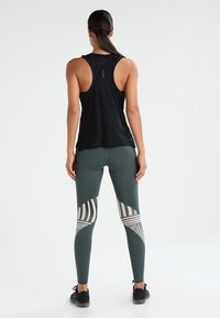 Donna che indossa un top senza maniche nero e leggings grigi con motivi geometrici in bianco e nero, in piedi di spalle su uno sfondo neutro.
