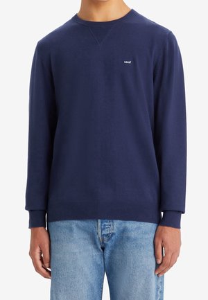 Mann trägt einen marineblauen Langarm-Levi's-Pullover kombiniert mit hellblauen Denim-Jeans, steht vor einem weißen Hintergrund.